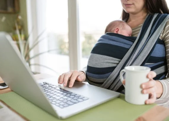 Mulher a usar echarpe de bebé às riscas azuis, cinza e pretas, com bebé ao colo, perto de computador portátil e caneca branca.