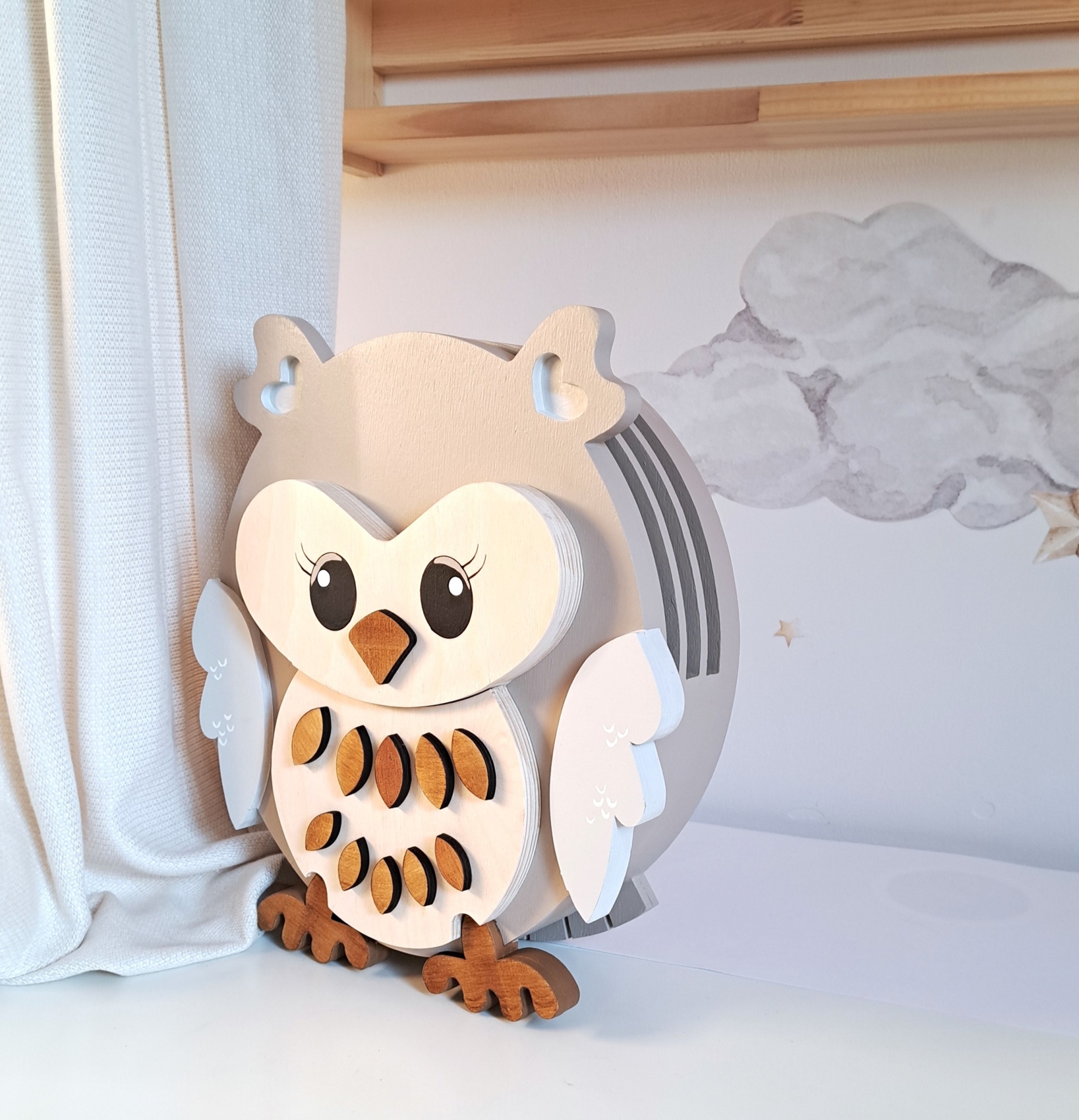 Candeeiro Coruja de Madeira Controlado por SMARTPHONE Abajur de madeira em forma de coruja com cores claras, olhos grandes e detalhes decorativos, numa mesa branca junto a cortinas e parede com nuvens desenhadas