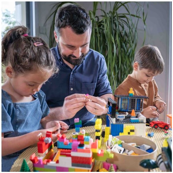 Base de Construção Infantil Compatível com LEGO® Adulto e crianças a construir com blocos de construção coloridos incluindo uma delegacia de polícia de brinquedo.