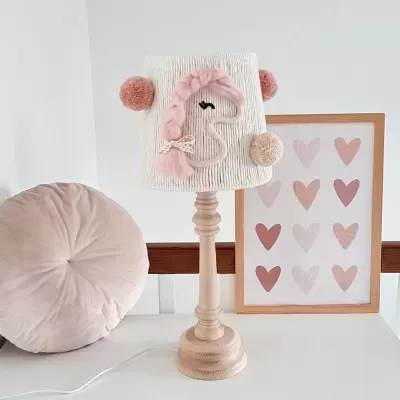 Candeeiro de mesa com abajur de unicórnio em tons de rosa ao lado de almofada redonda rosa e quadro com corações rosa