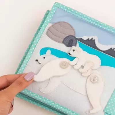 Livro infantil de tecido com ursos polares feitos de tecido e botões num cenário de montanhas e gelo