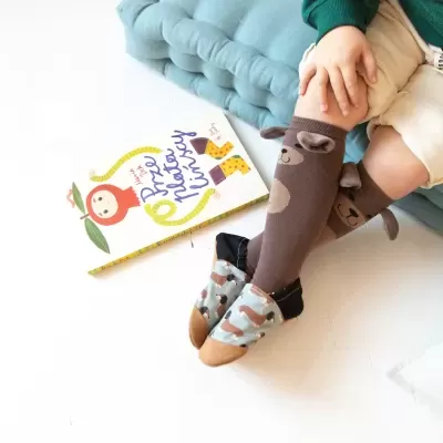 Meias de criança castanhas com urso, calçadas, criança sentada em tapete azul e livro colorido ao lado.