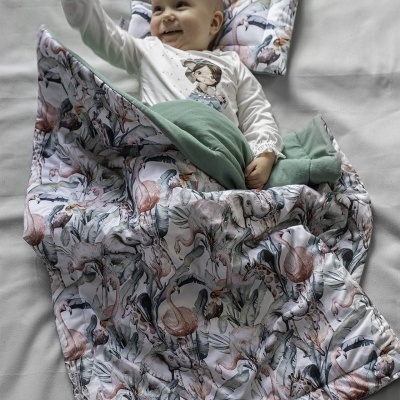 Bebé deitado sob edredão e almofada com padrão de aves, sorrindo e com pijama branco.