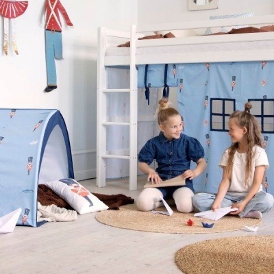 Cama alta com cortinas azuis e tenda infantil com padrão de guardas, crianças sentadas no tapete redondo no chão do quarto.