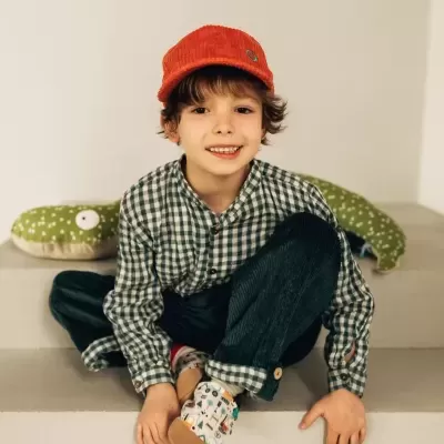 Menino sorridente com roupa casual e boné vermelho sentado em degraus brancos com peluche verde ao fundo.
