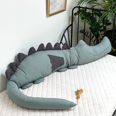 Almofada dinossauro azul esverdeado com espinhos cinzentos numa cama branca