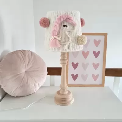 Candeeiro de mesa com base de madeira e abajur branco decorado com cavalo-marinho bordado, pompons e trança rosa junto a almofada e quadro de corações.