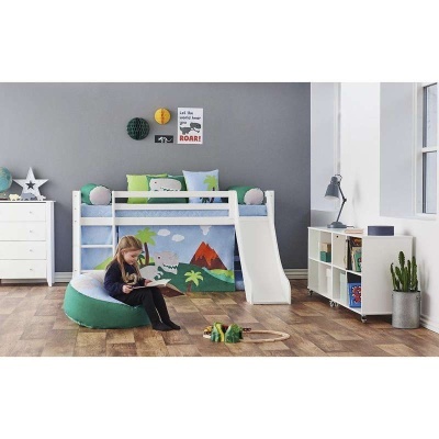 Quarto infantil com cama alta branca com escorrega, decoração de dinossauros, puff verde e branca, estante branca e criança a ler