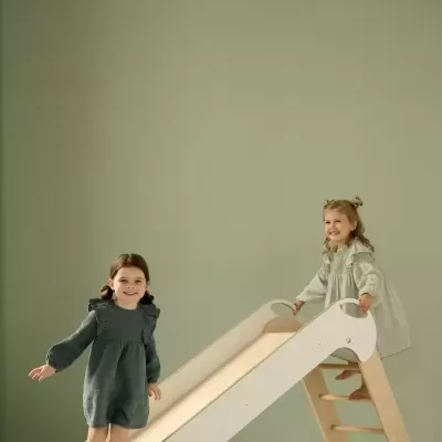 Duas crianças a brincar num escorrega branco com estrutura de madeira em fundo cinzento.