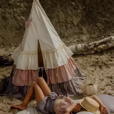 Tenda tipi infantil com tecido creme, rosa e cinza em praia de areia, menina de chapéu de palha deitada ao lado
