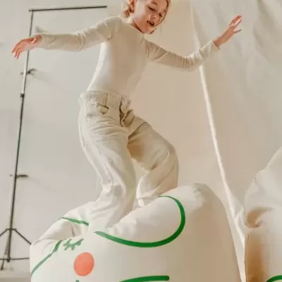 Menina com roupa clara em cima de puff branco com padrão verde e laranja num espaço interior claro