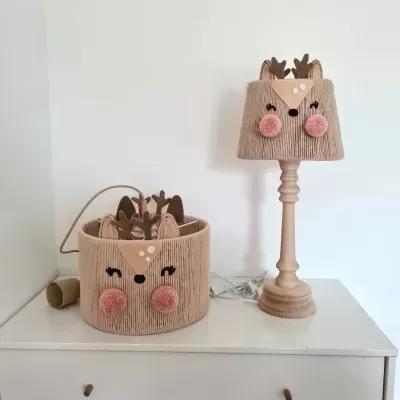 Candeeiro de mesa e cesto redondo com rosto de rena em tecido bege decorativos sobre uma cómoda branca