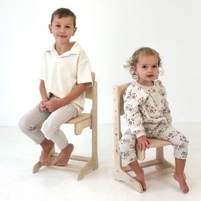 Crianças sentadas em cadeiras de madeira, roupas claras, fundo branco.