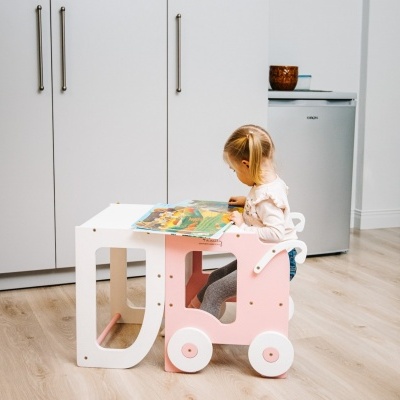 Criança sentada numa cadeira rosa a ler um livro numa mesa branca em ambiente interior