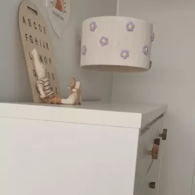 Abajur branco com flores roxas suspenso sobre móvel branco com placa de madeira do alfabeto.