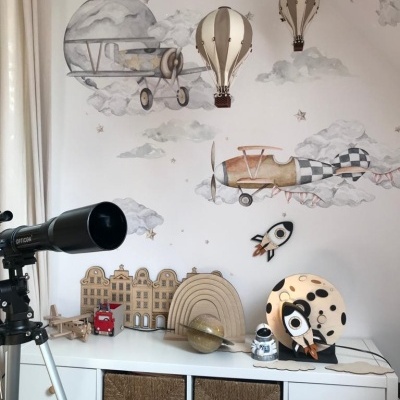 Decoração infantil com tema de aviões e espaço, móveis brancos, brinquedos de madeira e telescópio preto
