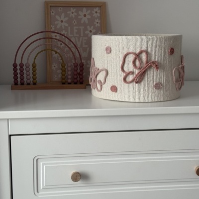 Abajur branco com borboletas rosa sob cómoda branca, com moldura e arco-íris decorativo ao fundo