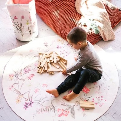 Criança a brincar com blocos de madeira sobre tapete redondo floral, saco de tecido e cobertores ao lado