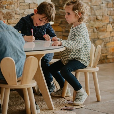 Mesa redonda branca com cadeiras de madeira clara em ambiente com parede de pedra, crianças sentadas à mesa