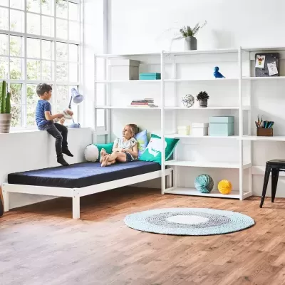 Quarto infantil com cama branca, colchão azul, estante branca, e tapete redondo azul e cinzento em chão de madeira clara