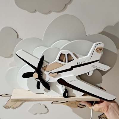 Avião de madeira branco, bege e preto com hélice sobre fundo de nuvens decorativas.