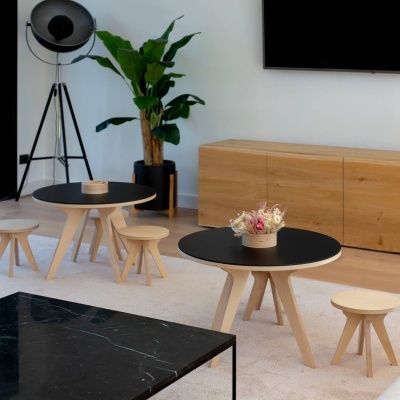 Salão com mesas redondas com tampo preto e bancos pequenos de madeira clara, móvel baixo de madeira, planta e candeeiro de pé.