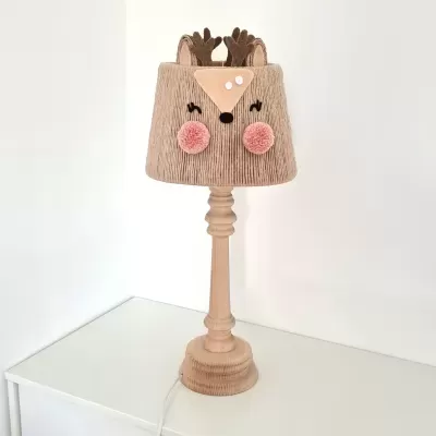 Candeeiro de mesa com sombra em forma de veado em tecido castanho claro e base em madeira clara sobre mesa branca