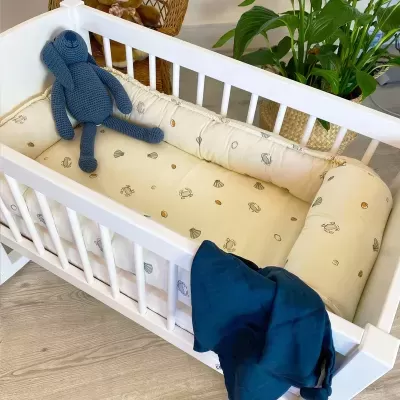 Berço branco de madeira com colchão e almofadas bege com padrão de mar e boneco de tricô azul