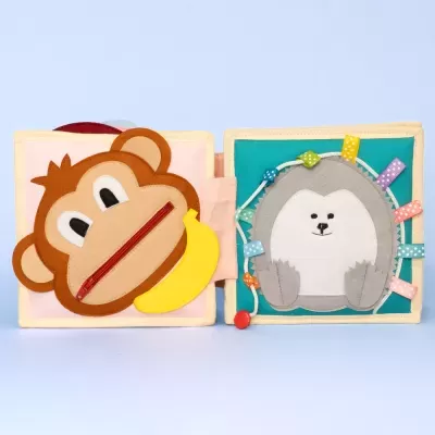 Livro infantil de tecido com capa de macaco e página interior com rosto de animal e fitas coloridas.