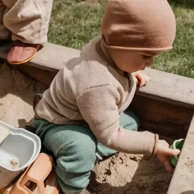 Criança com roupas de malha e gorro rosa brincando numa caixa de areia com brinquedos.
