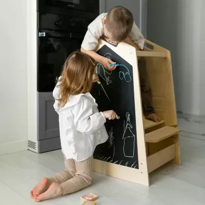 Cavalete educativo em madeira com quadro negro onde duas crianças desenham com giz colorido.