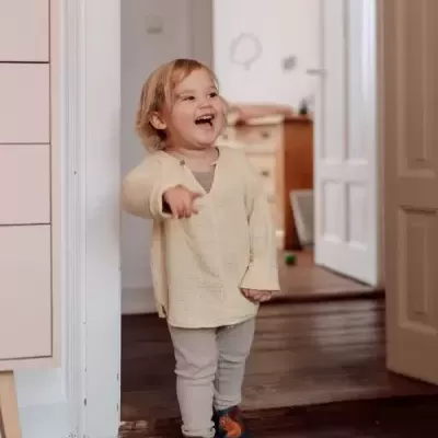 Criança sorridente vestindo camisola creme, calças cinza e sapatos azuis com padrão laranja