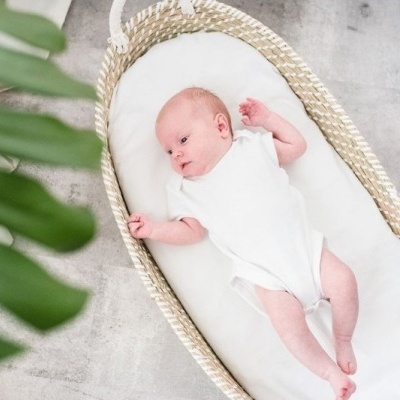 Berço oval de palha com colchão branco e bebé vestido de branco em chão cinzento