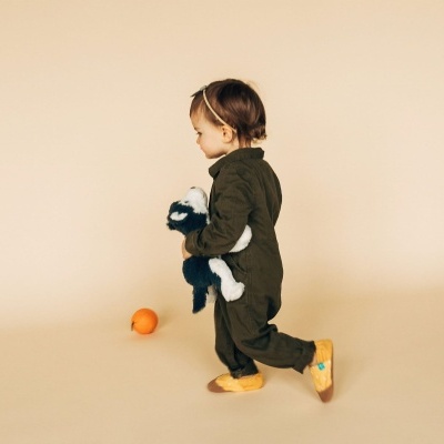 Criança com macacão verde escuro e peluche segurando uma tangerina no chão