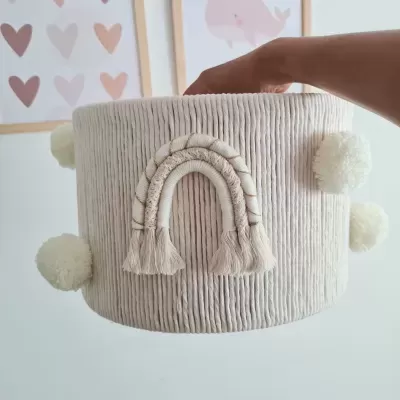 Cesto redondo cinza claro com pompons e arco-íris bordado segurado por uma mão