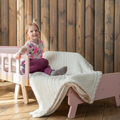 Cama infantil de madeira rosa claro com manta branca e criança sentada.