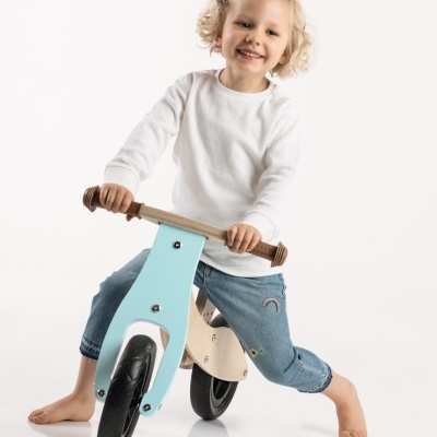 Criança numa bicicleta de equilíbrio de madeira com parte frontal azul clara e punhos castanhos, sobre fundo branco