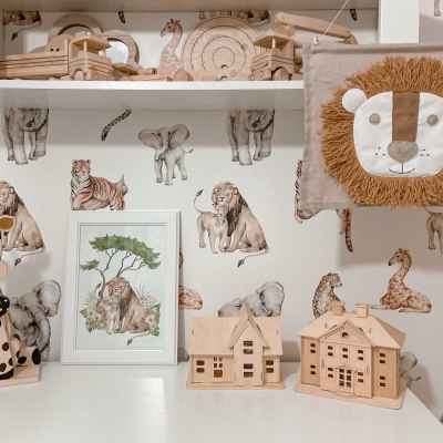 Conjunto de brinquedos de madeira e decoração infantil com tema de animais, incluindo prédios, quadro de leões, boneco girafa e almofada de leão.