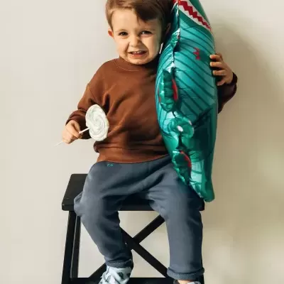 Criança com camisola castanha e calças azuis segurando balão em forma de tubarão e pirulito branco sentado num banco preto.