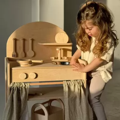 criança brincando com cozinha de madeira para crianças com utensílios e cortinas xadrez.