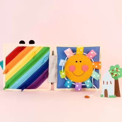 Livro de tecido educativo colorido com zíperes e sol sorridente em fundo cor-de-rosa.