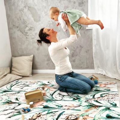 Tapete decorativo infantil com padrão floral, mulher segurando bebé vestido de verde sobre o tapete