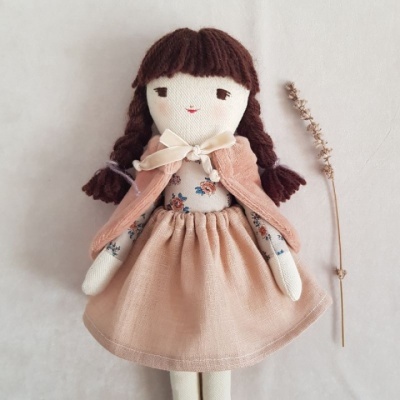 Boneca de pano vestida com vestido rosa e capa, com tranças castanhas, colocada ao lado de um ramo seco sobre fundo claro