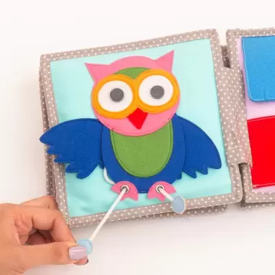 Livro de tecido colorido para crianças com coruja azul, verde, rosa e amarelo e bordas cinza com bolinhas brancas.