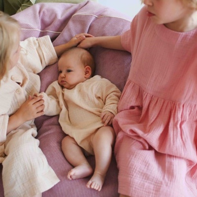 Três crianças vestindo roupas creme e rosa sentadas em tecido roxo com textura riscada