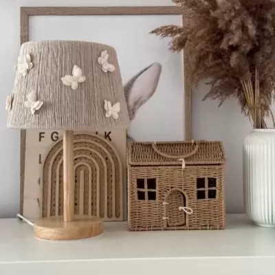 Abajur de madeira com abajur bege decorado, caixa de transporte em forma de casa em material trançado, flores secas em vaso branco