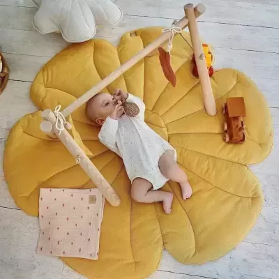 Bebé deitado num tapete almofadado amarelo em formato de folha com estrutura de brincar de madeira