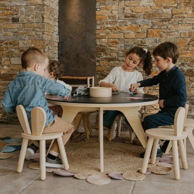 Mesa redonda preta com quatro cadeiras de madeira clara com encostos em forma de orelhas de coelho e quatro crianças sentadas