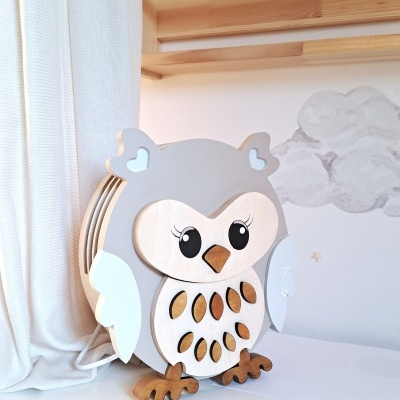 Abajur coruja de madeira com cores cinza e branco, sobre superfície branca junto a cortina e parede com nuvens