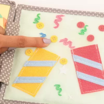 Livro sensorial de tecido com elementos coloridos e texturizados em páginas verdes e capa cinzenta com bolinhas.
